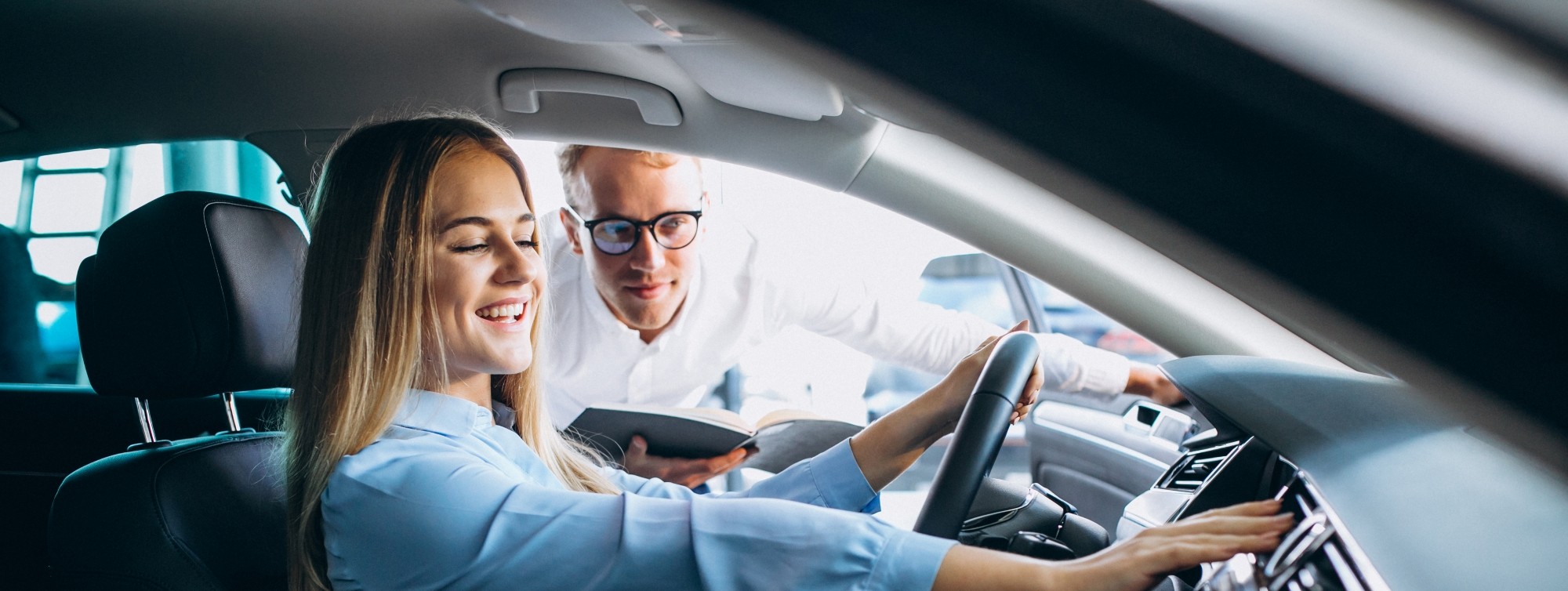A smiling woman sitting in the driver's seat of a car while a salesman looks in from the window.